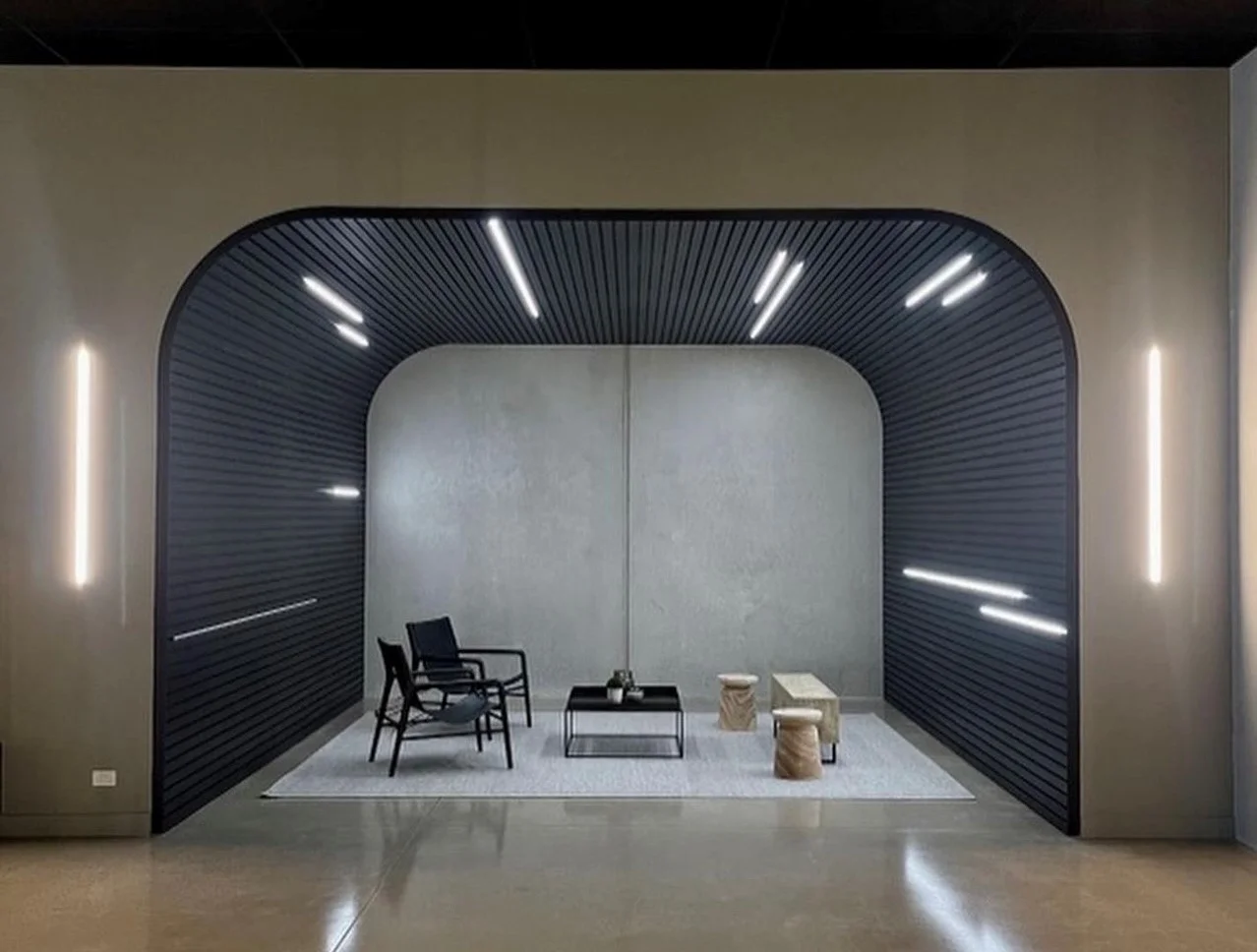 A modern lounge area features black ribbed arch walls with integrated LED lights, minimalist black chairs, a black coffee table, and two wooden stools, all set on a light gray rug against a plain gray background.