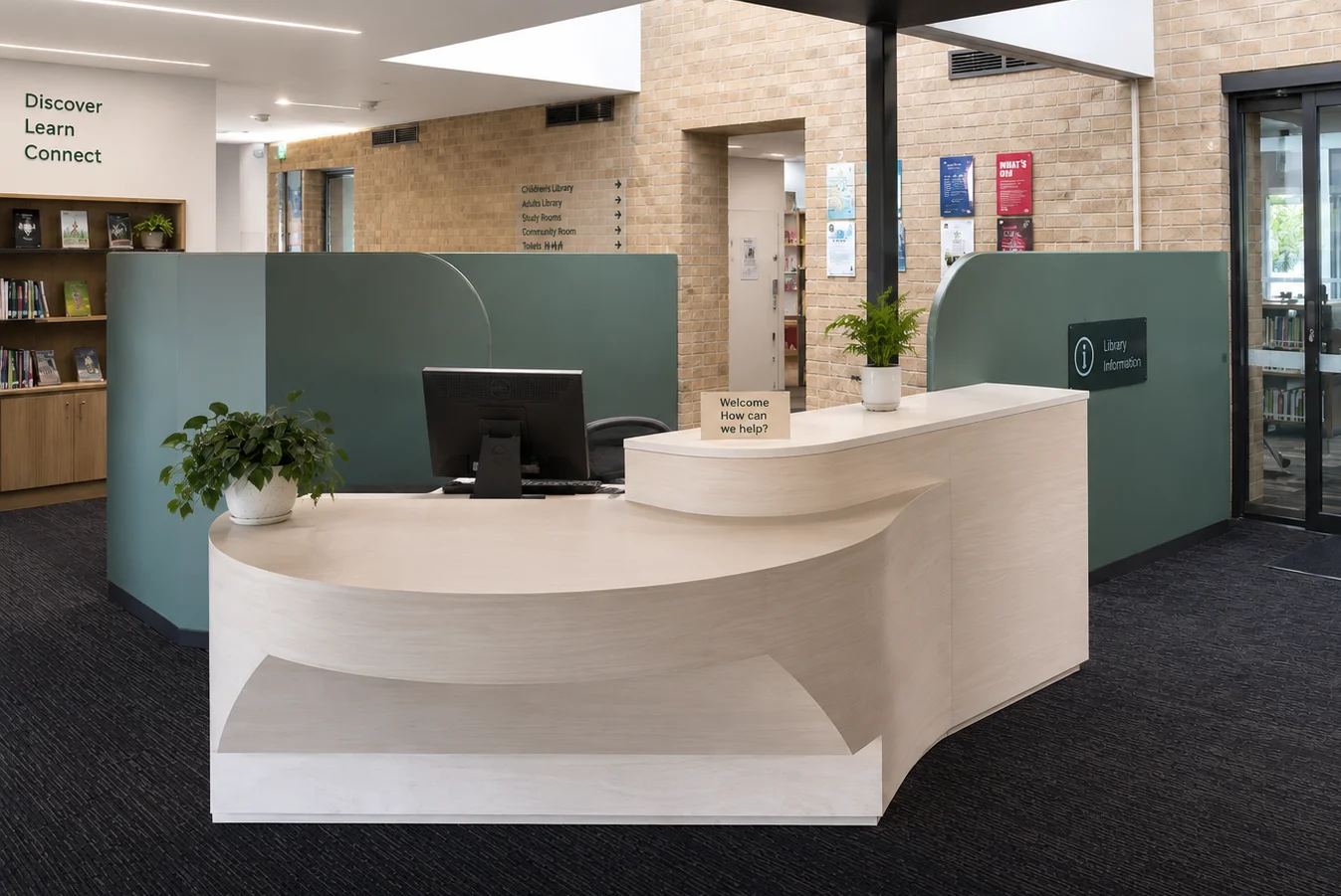 Modern library reception area with a curved white desk, computer monitor, potted plants, and a small "Welcome" sign. Shelves of books and informational signs are visible in the background.