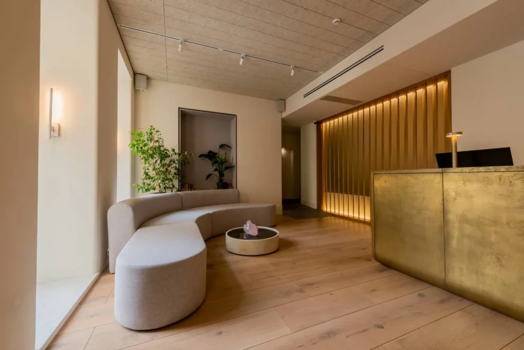 Modern reception area with a curved beige sofa, round coffee table, potted plants, a golden front desk with two monitors, wood flooring, and warm lighting from wall sconces and ceiling fixtures.