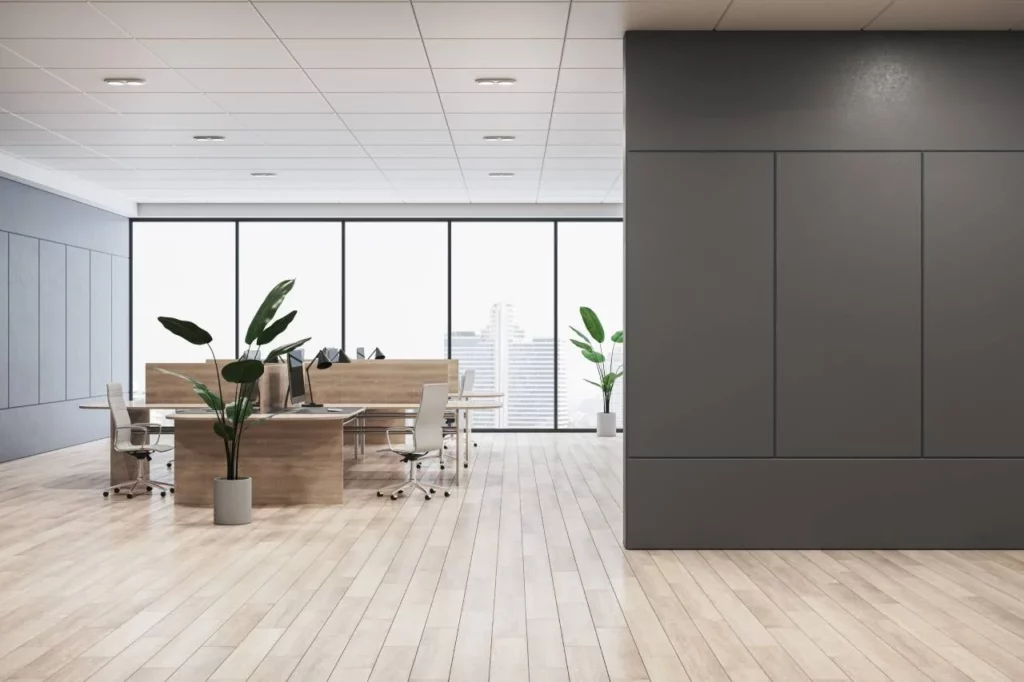 Modern office space with wooden floors, large windows, several desks with computers and chairs, and potted plants. The room is bright and spacious with neutral tones and a city view outside the windows.