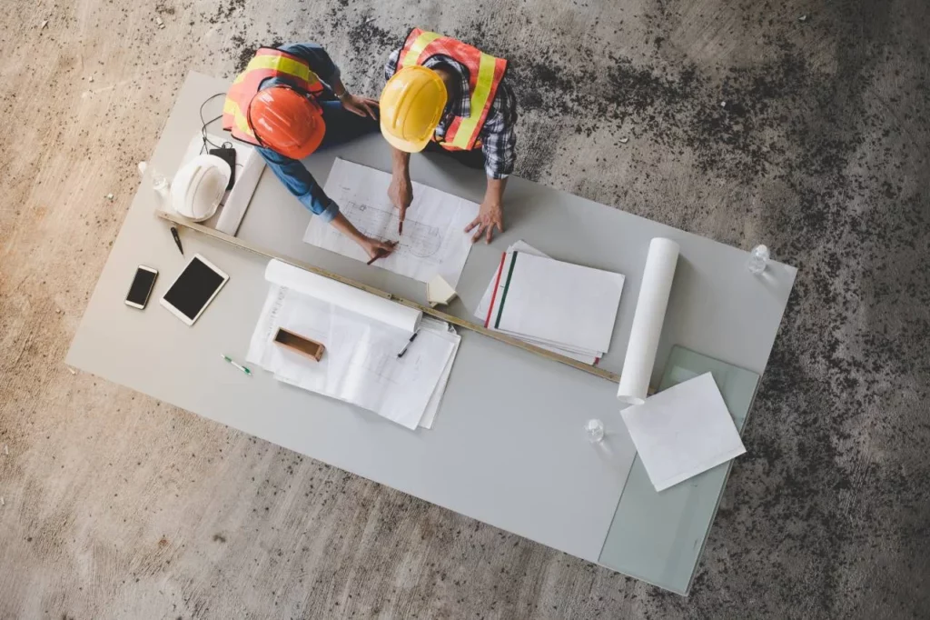 Two construction workers wearing safety vests and helmets review blueprints on a large table with documents, a hard hat, tablet, and glass of water, viewed from above.