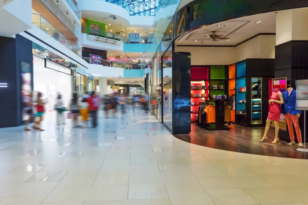 A brightly lit indoor shopping mall with several floors, blurred shoppers walking, and a colorful clothing store with mannequins dressed in stylish outfits near the right side of the image.