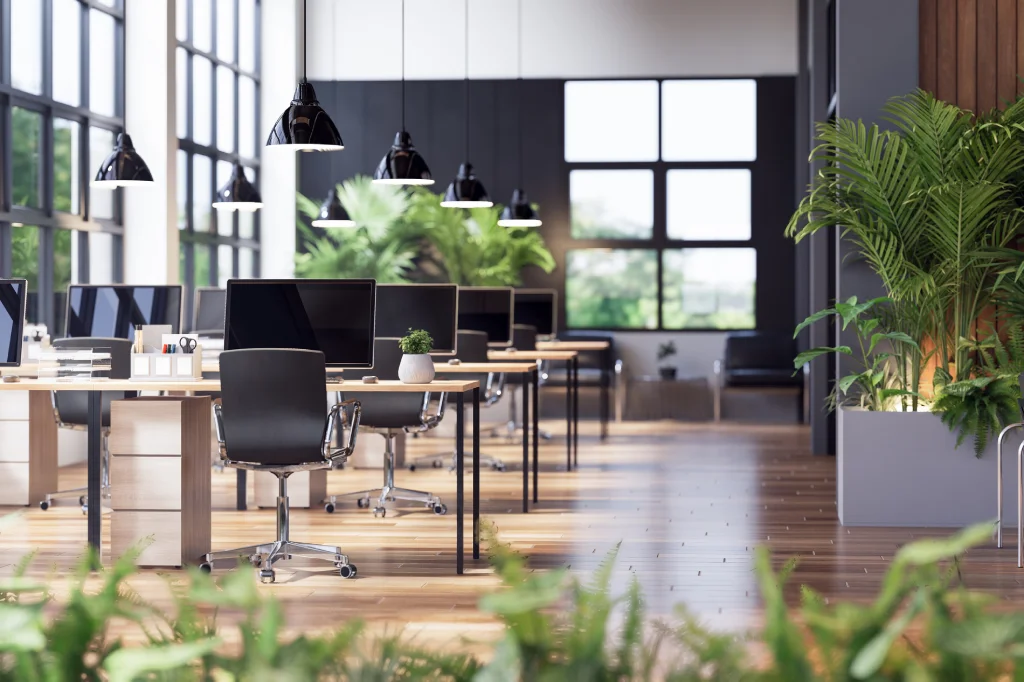 Modern open-plan office with rows of desks, black chairs, computers, and potted plants; large windows let in natural light, and hanging black lamps add to the bright, green, and inviting workspace atmosphere.