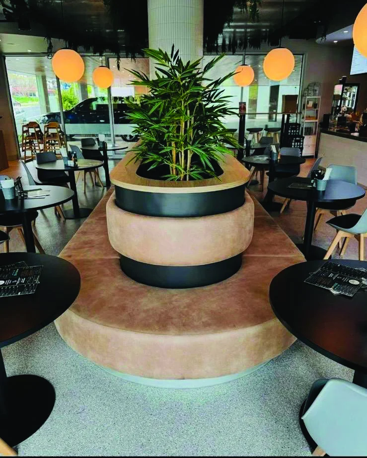 A modern cafe interior with round tables, black chairs, and a large circular seating area featuring a central planter with green plants. Warm pendant lights hang from the ceiling, creating a cozy atmosphere.