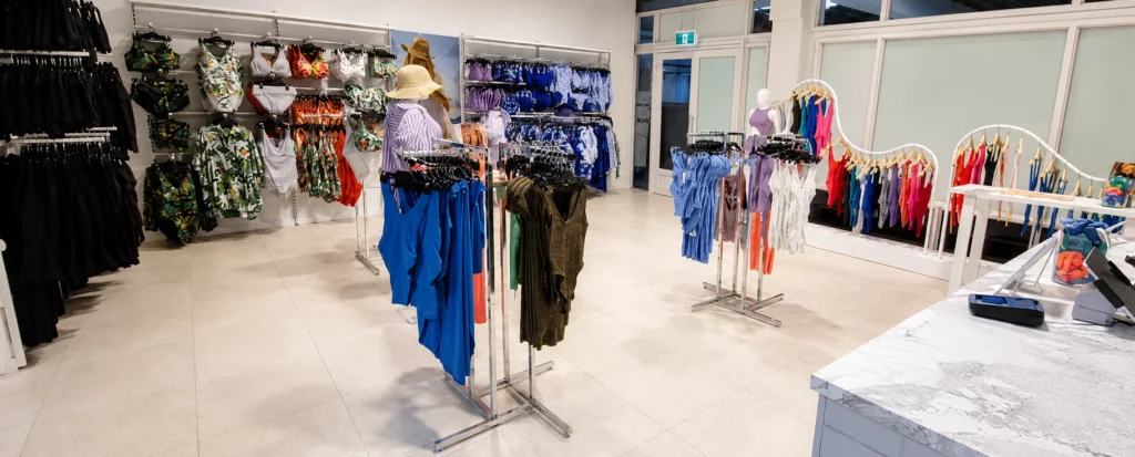 A brightly lit swimwear store with racks displaying colorful swimsuits, bikinis, and cover-ups. Sun hats are on display, and the counter is visible in the foreground. The store has a clean, modern design.