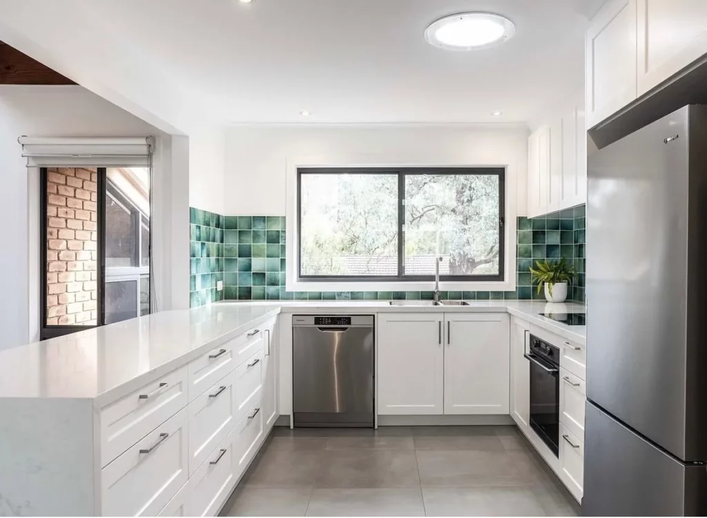 Modern kitchen with white cabinets, stainless steel appliances, green tiled backsplash, large window above the sink, and potted plant on the counter. Natural light brightens the space.